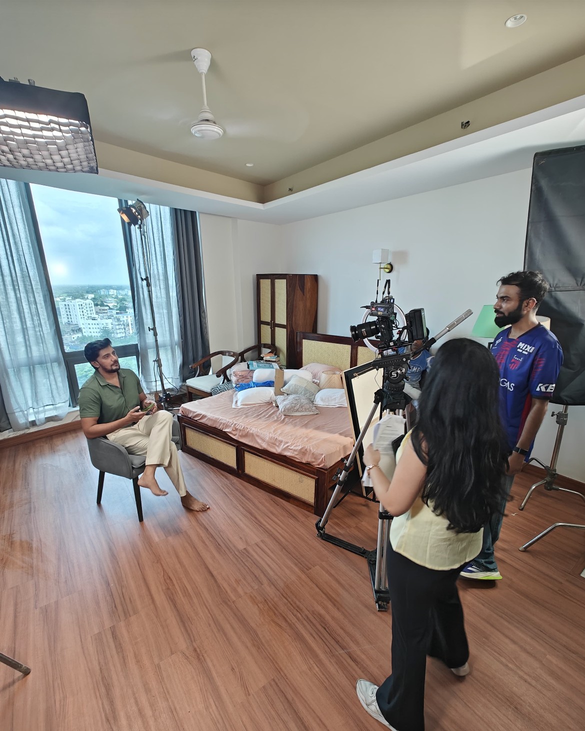 Behind the scenes of a commercial shoot: an interviewee seated in an armchair while a camera operator films him and a crew member holds cue scripts.